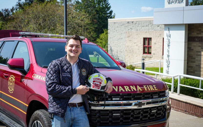 A dark haired man stands in front of the Baker Institute wearing a Varna firefighter branded jacket. 