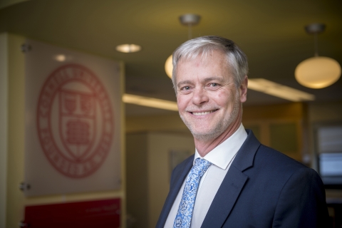 A headshot showing a man standing in the Baker Lobby with the Cornell seal behind him. 