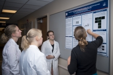 A group of scientists gather around a person in a black shirt at a corkboard. 