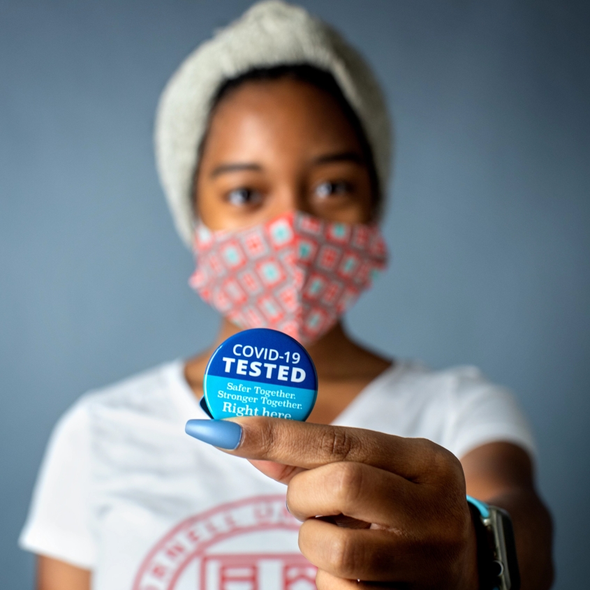 Woman wearing a mask and holding a pin that says COVID-19 Tested