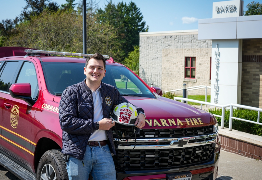 A dark haired man stands in front of the Baker Institute wearing a Varna firefighter branded jacket. 