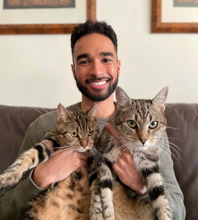 A man with dark short hair holds two striped cats while sitting on a couch. 