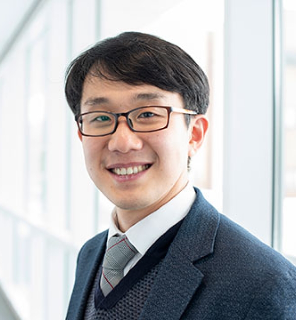 Esak Lee in a grey suit and wearing black glasses while standing in front of a wall of windows.