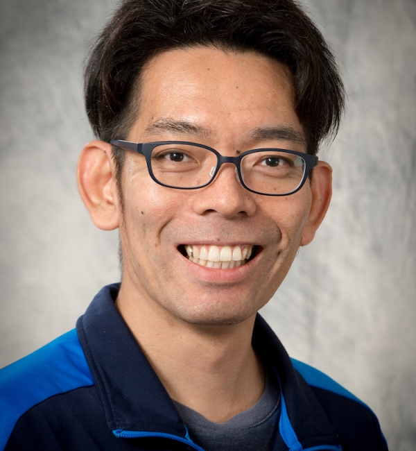 Toshi Kawate in a grey background with a blue-lined black windbreaker and black glasses.